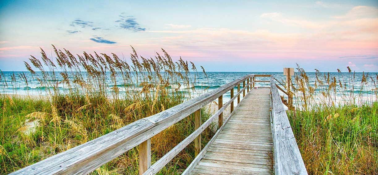 Photograph of a narrow old wooden pier stretching out to a beach and wave churning ocean.