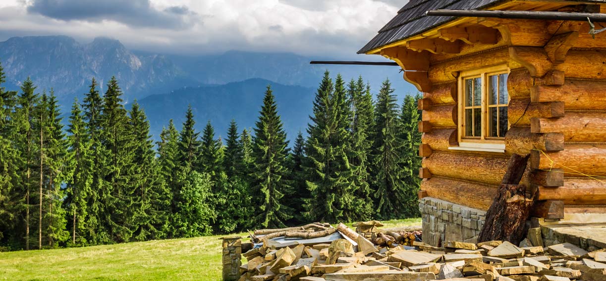 A cabin in the mountains beyond is fur trees and mountains capped with white & gray clouds.